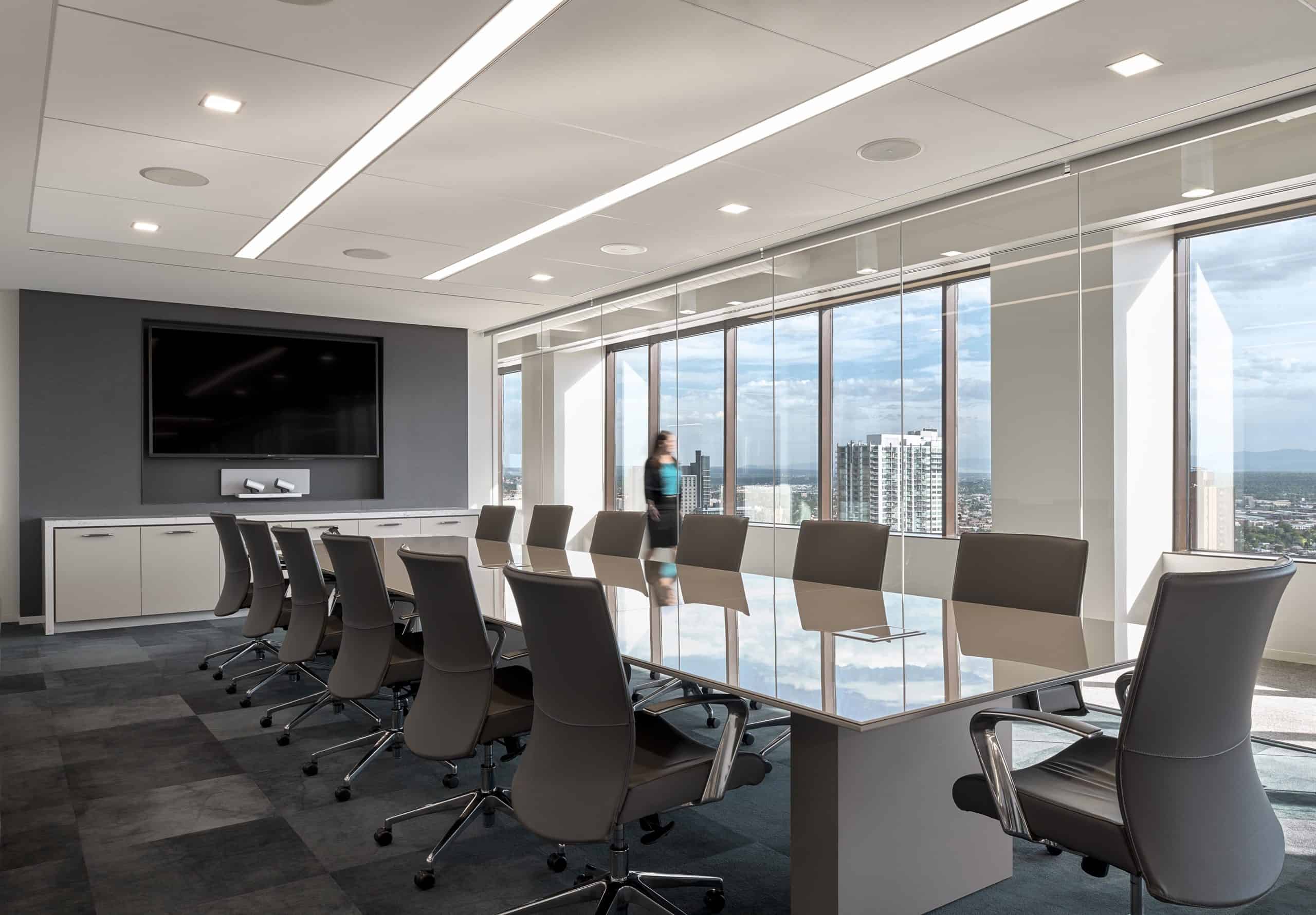 Modern room: glass table, gray chairs, screen on wall. Large windows view skyscrapers. Person at window, cloudy sky outside.